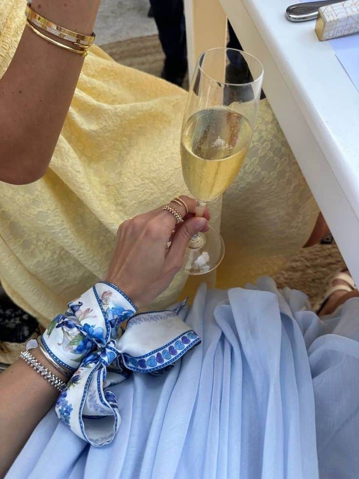 _ 46 Woman toasting with champagne in elegant floral attire at a stylish event, showcasing jewelry and a blue scarf with intricate design. | Sky Rye Design Woman toasting with champagne in elegant floral attire at a stylish event, showcasing jewelry and a blue scarf with intricate design.