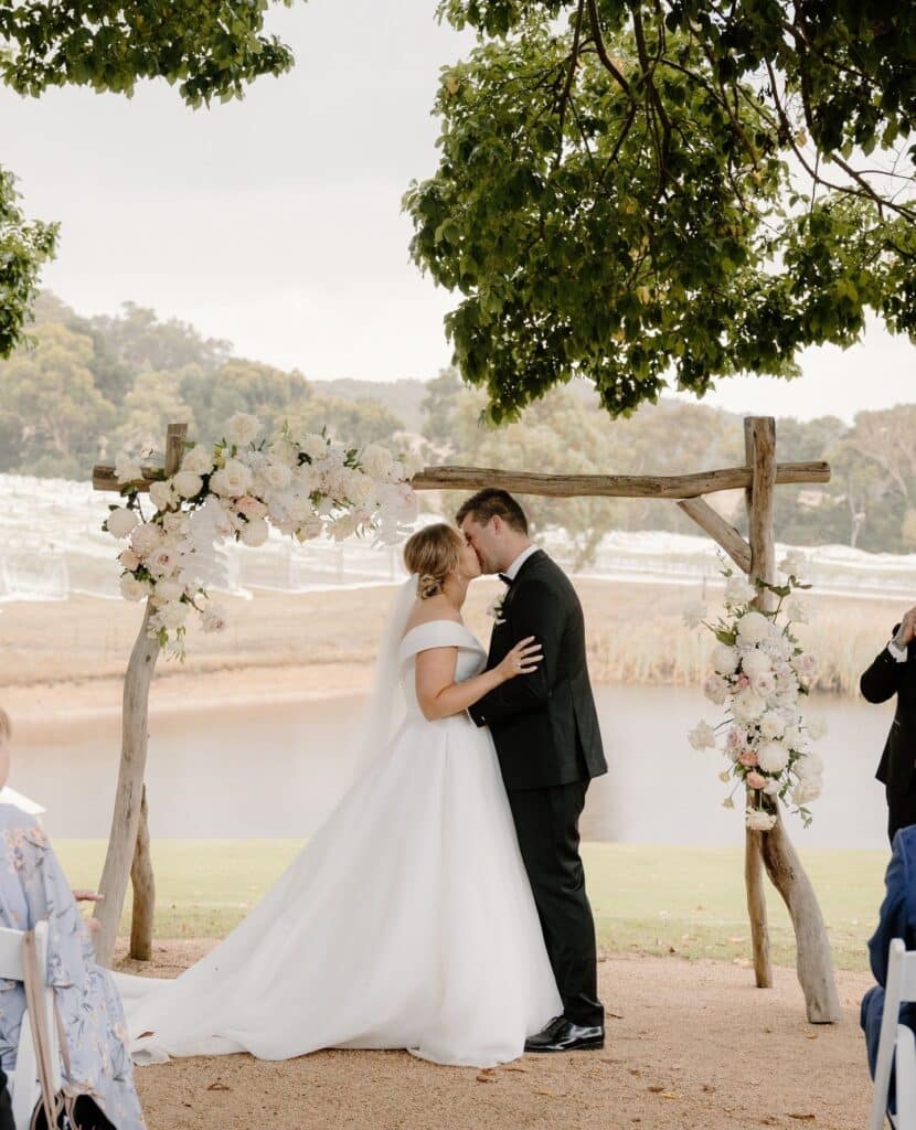 464182166_2797004990467286_8331027381878737131_n Bride and groom share a kiss under a rustic floral arch outdoors, surrounded by lush greenery and a serene lake backdrop. | Sky Rye Design Bride and groom share a kiss under a rustic floral arch outdoors, surrounded by lush greenery and a serene lake backdrop.
