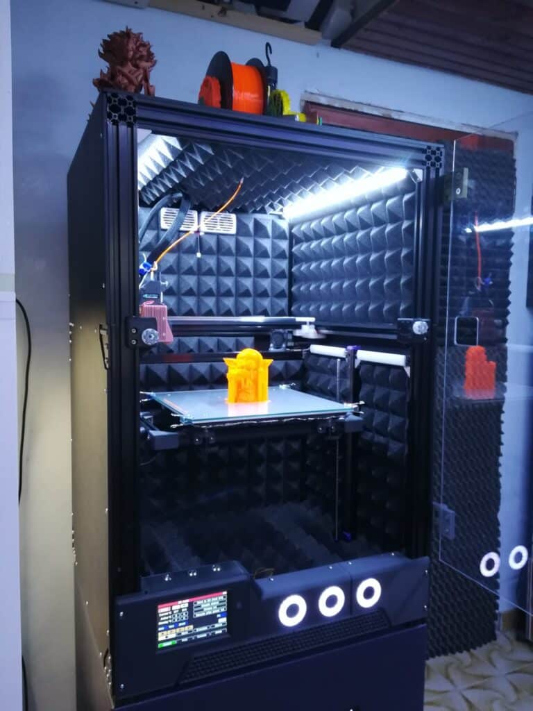 3D printer with orange filament and acoustic foam enclosure creating a detailed orange model, under LED lighting in a workshop.
