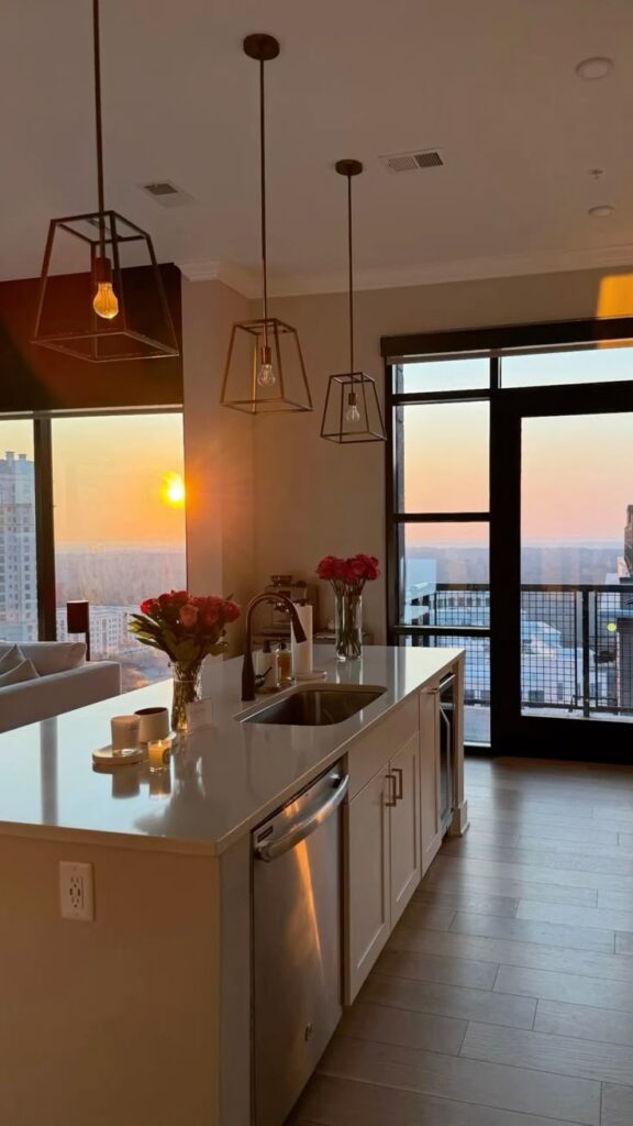 Modern kitchen with island, pendant lights, and city view at sunset through large windows. Cozy ambiance with flowers and candles.