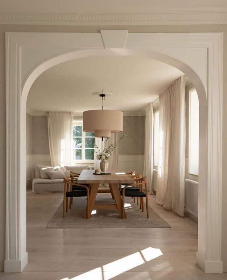 Modern dining room with wooden table, chairs, and soft beige tones. Arched entrance, large window, and elegant lighting. Cozy and inviting.