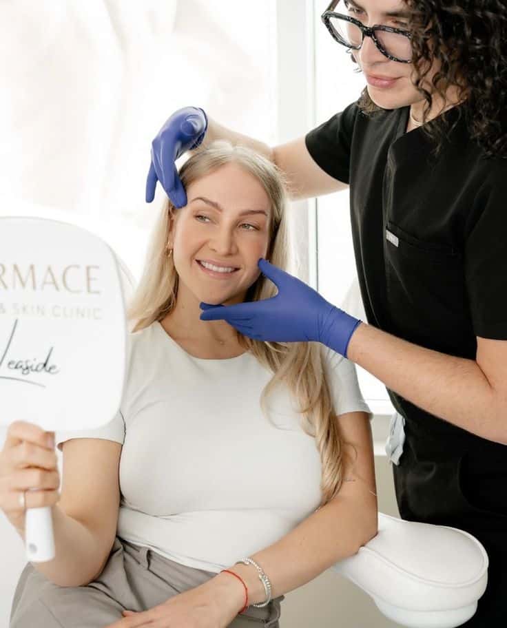 _ 5 Woman receiving skincare consultation at clinic, smiling at aesthetician with gloves, holding mirror. | Sky Rye Design Woman receiving skincare consultation at clinic, smiling at aesthetician with gloves, holding mirror.