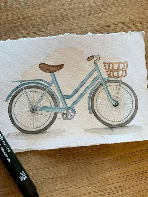_ 5 Watercolor illustration of a vintage blue bicycle with a front basket on textured paper, resting on a wooden surface. | Sky Rye Design Watercolor illustration of a vintage blue bicycle with a front basket on textured paper, resting on a wooden surface.