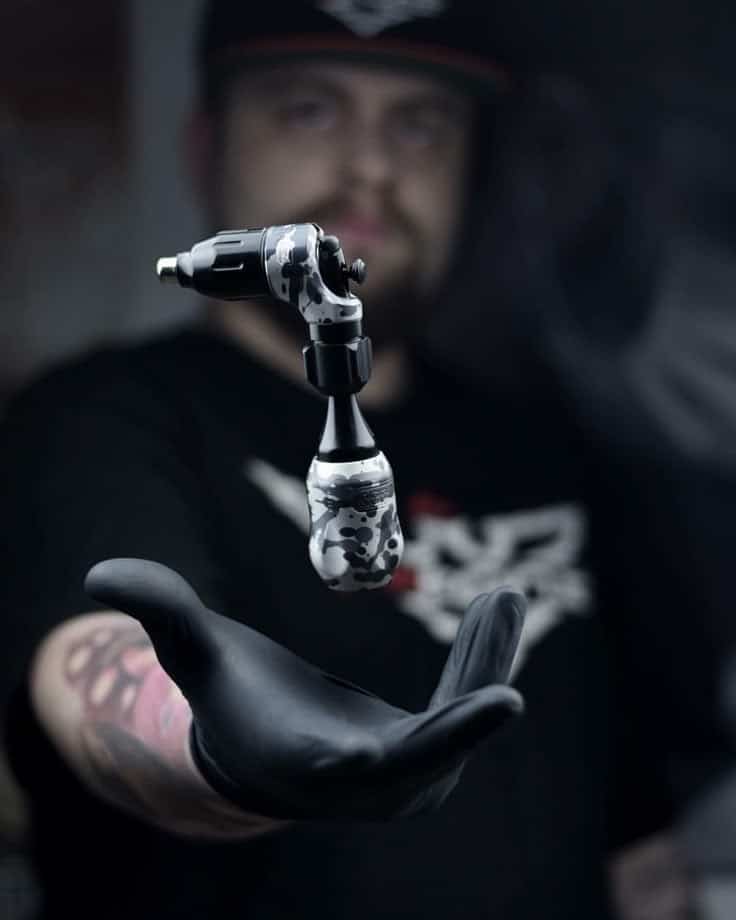 _ 5 Tattoo artist holding black and white tattoo machine in hand with dark glove, demonstrating precision equipment in studio setting. | Sky Rye Design Tattoo artist holding black and white tattoo machine in hand with dark glove, demonstrating precision equipment in studio setting.