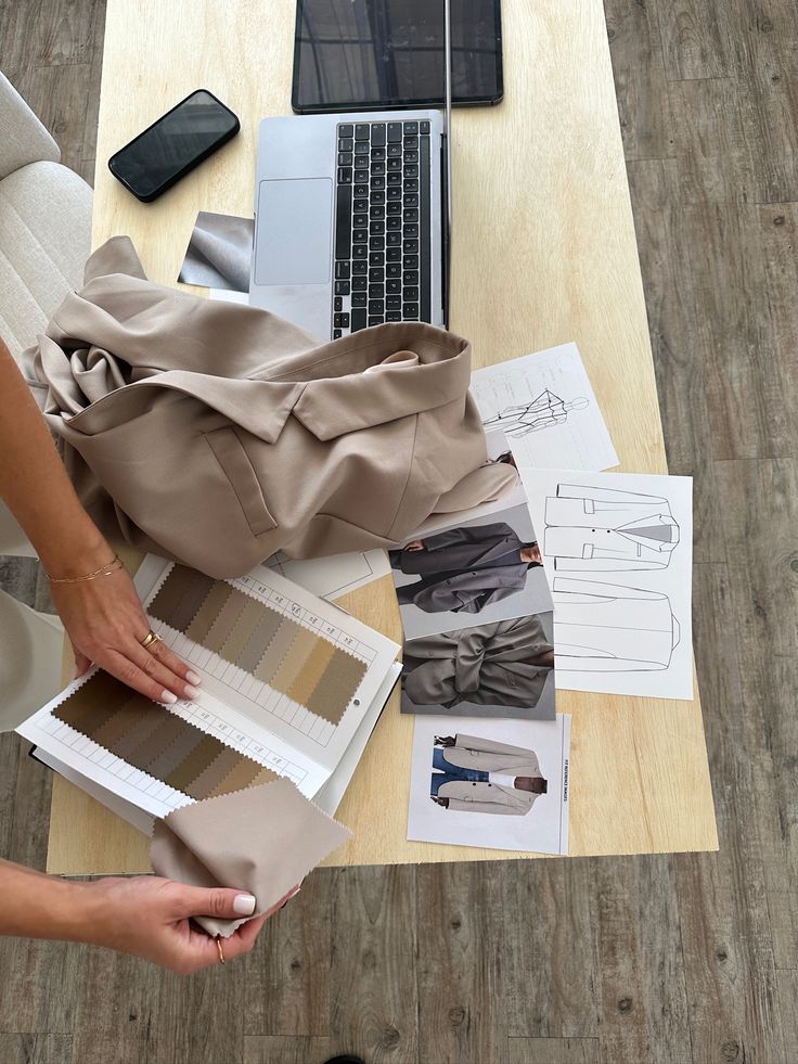 Designer planning outfit with fabric swatches and sketches on a desk with a laptop and phone, focusing on fashion coordination.
