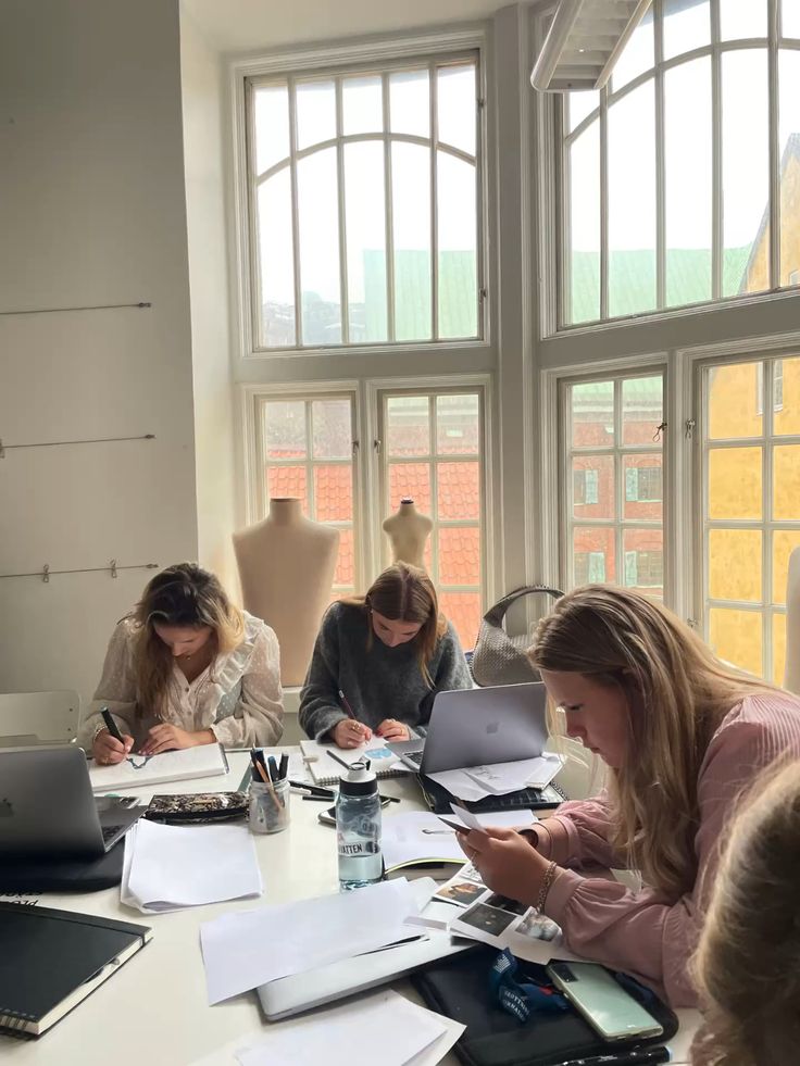 _ 5 Fashion students working on designs at a table with laptops, papers, and mannequins in a sunlit room. | Sky Rye Design Fashion students working on designs at a table with laptops, papers, and mannequins in a sunlit room.