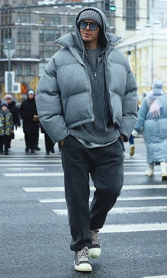 Man in stylish winter outfit crossing street, wearing gray puffer jacket, knit beanie, sunglasses, and sneakers, in urban setting.
