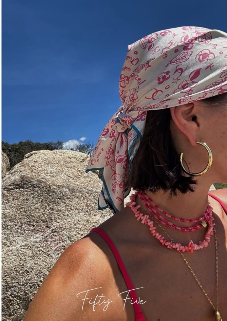 _ 51 Woman wearing a patterned scarf, hoop earring, and layered necklaces at the beach under a clear blue sky. | Sky Rye Design Woman wearing a patterned scarf, hoop earring, and layered necklaces at the beach under a clear blue sky.