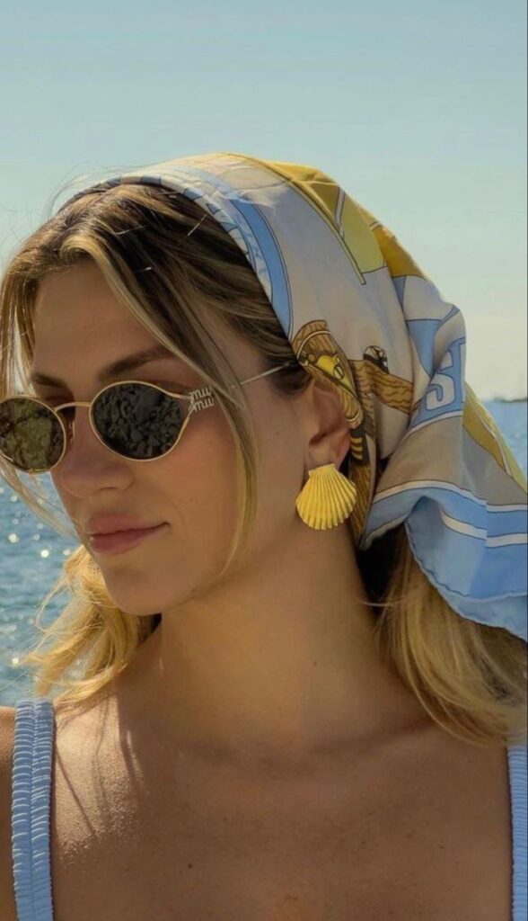 _ 52 Woman at the beach wearing sunglasses, a patterned headscarf, and shell earrings, enjoying a sunny day by the sea. Fashionable summer style. | Sky Rye Design Woman at the beach wearing sunglasses, a patterned headscarf, and shell earrings, enjoying a sunny day by the sea. Fashionable summer style.