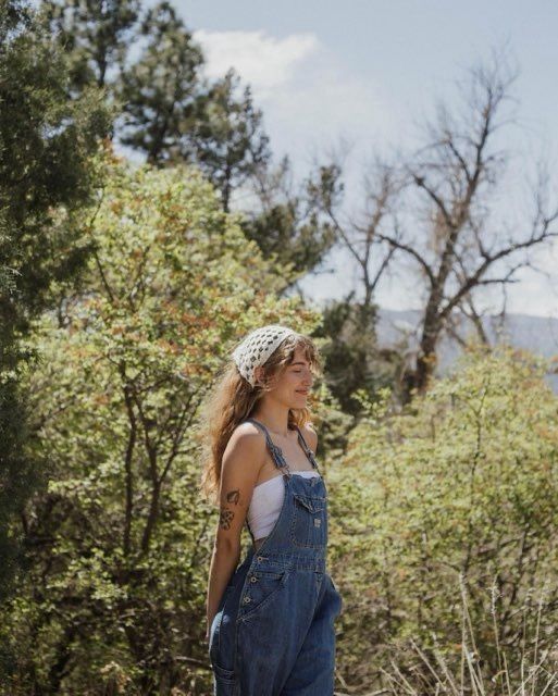 _ 53 Woman in denim overalls with a headscarf stands in a lush forest, basking in the sunlight on a clear day. | Sky Rye Design Woman in denim overalls with a headscarf stands in a lush forest, basking in the sunlight on a clear day.
