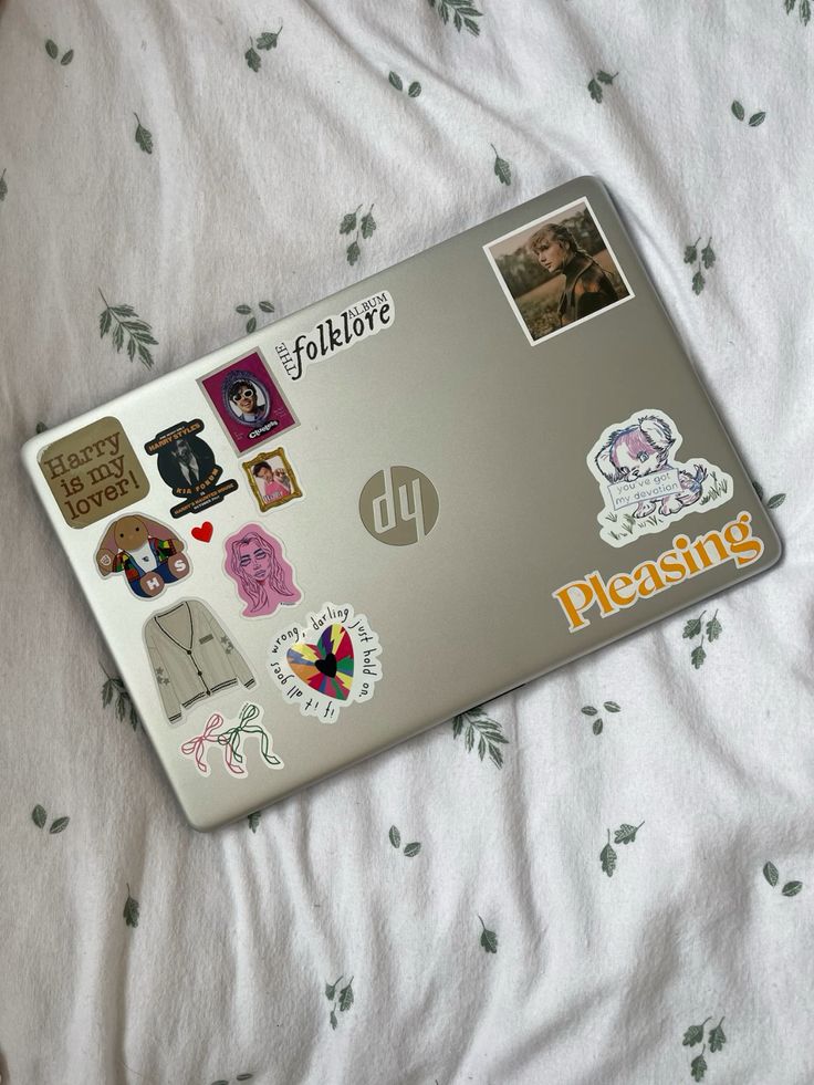 _ 59 Laptop adorned with pop culture and music-themed stickers on a leaf-patterned bedspread. | Sky Rye Design Laptop adorned with pop culture and music-themed stickers on a leaf-patterned bedspread.