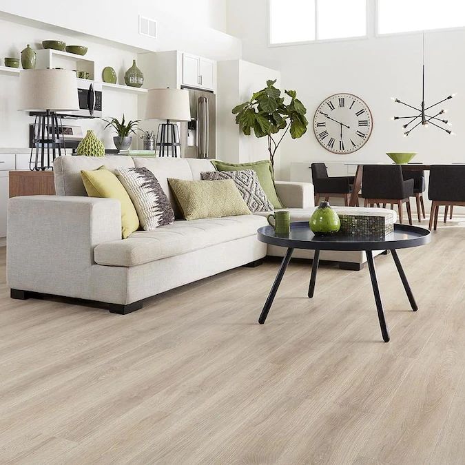 Modern living room with beige sofa, green accents, round black table, and wood flooring. Open dining area in background.