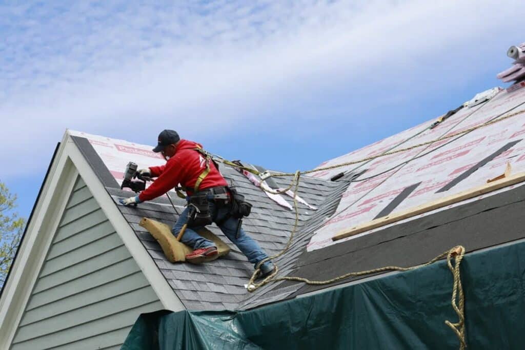 Roofer in safety gear installs shingles on a residential roof under clear skies, ensuring durable home protection.