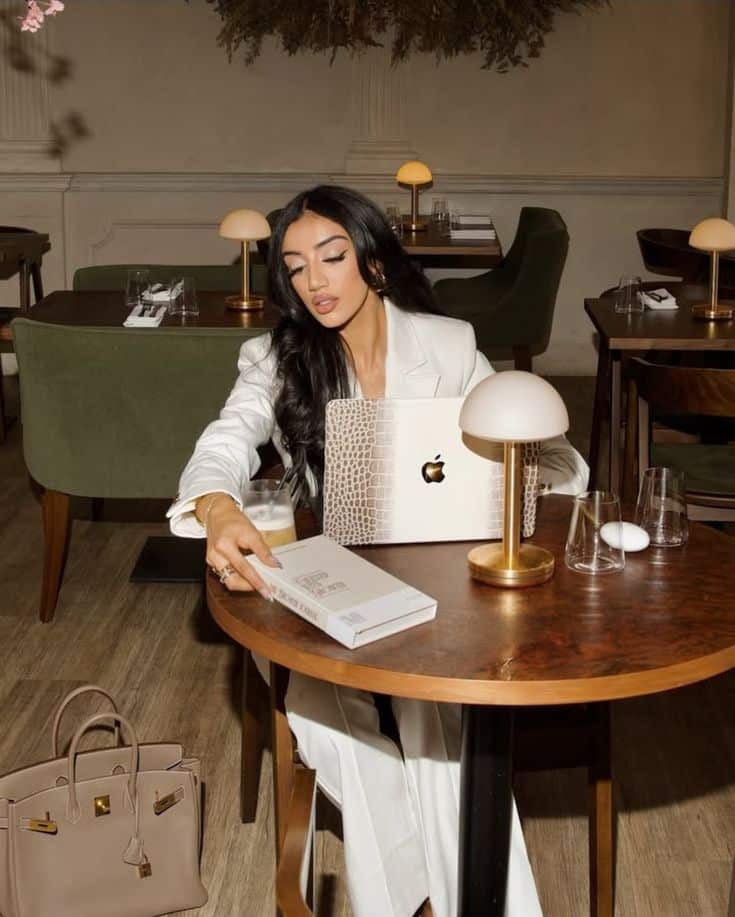 Woman in a chic cafe working on a luxury laptop, with a designer bag on the floor, creating a stylish, modern workspace vibe.