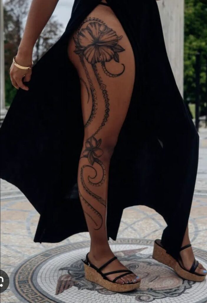 _ 6 Woman in black dress showing elegant floral leg tattoo, standing on detailed mosaic floor. | Sky Rye Design Woman in black dress showing elegant floral leg tattoo, standing on detailed mosaic floor.