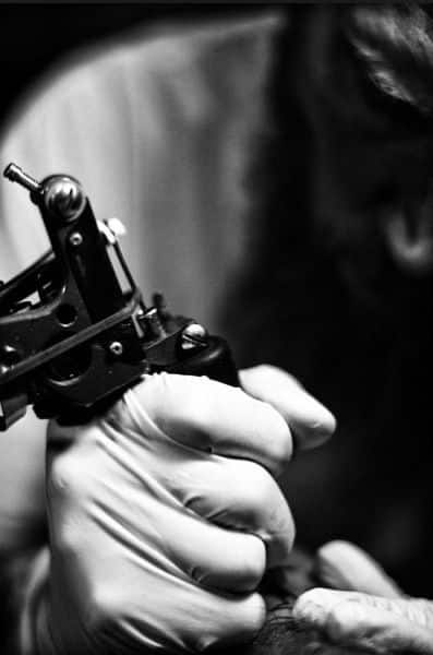 _ 6 Tattoo artist in gloves holding a tattoo machine, focused on detail work in black and white. | Sky Rye Design Tattoo artist in gloves holding a tattoo machine, focused on detail work in black and white.