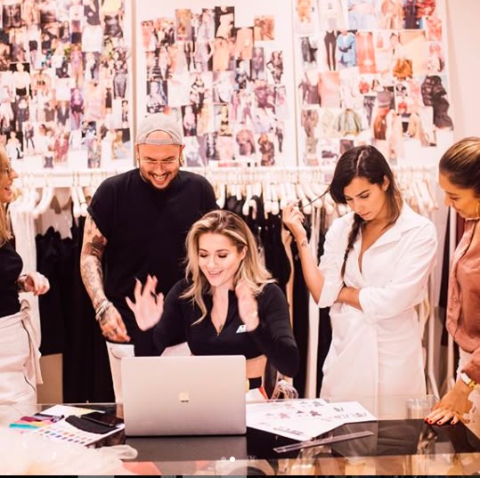 Team collaborating on a fashion project around a laptop, design sketches visible, mood boards in the background, fashion studio setting.