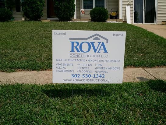 Rova Construction LLC sign on a lawn, offering services like remodeling, carpentry, and flooring, with contact info and website.