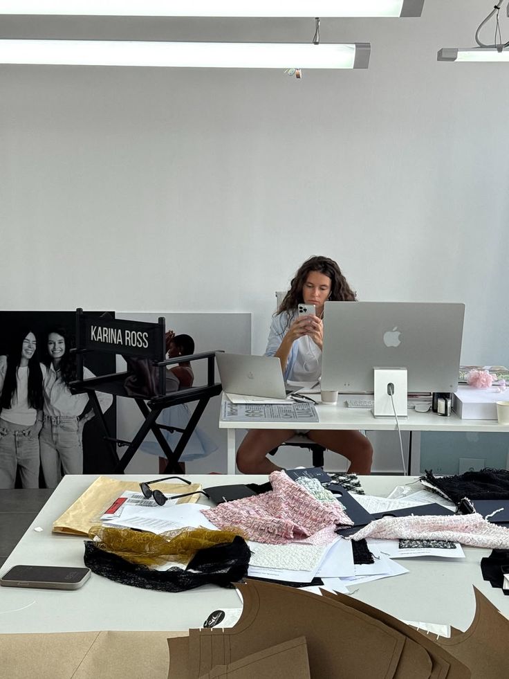 Designer working on a project at a modern office desk, surrounded by fabrics and sketches, capturing the moment on a smartphone.