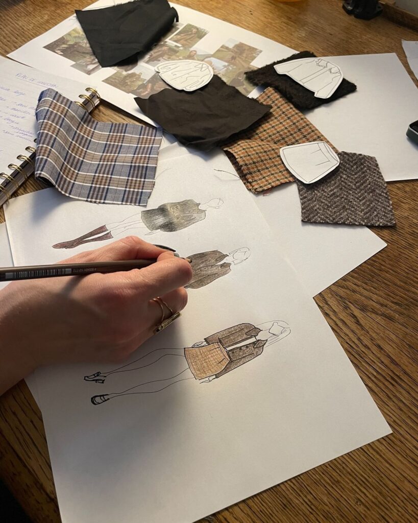 604c7a7dde1b9acd19c5546fcf37b3e8 Fashion designer sketching a clothing design with fabric swatches on a wooden table, focusing on plaid and textured patterns. | Sky Rye Design Fashion designer sketching a clothing design with fabric swatches on a wooden table, focusing on plaid and textured patterns.