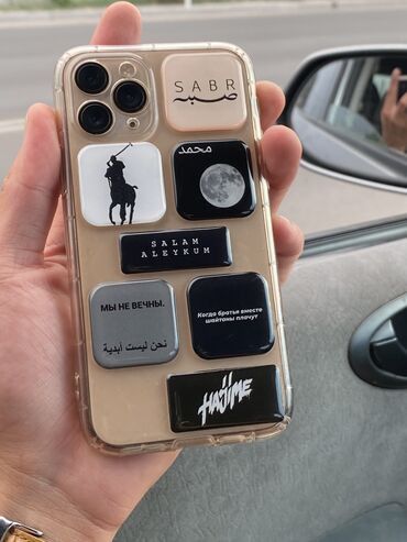 _ 63 Person holding a smartphone with a clear case featuring various graphic and text stickers near a car interior. | Sky Rye Design Person holding a smartphone with a clear case featuring various graphic and text stickers near a car interior.