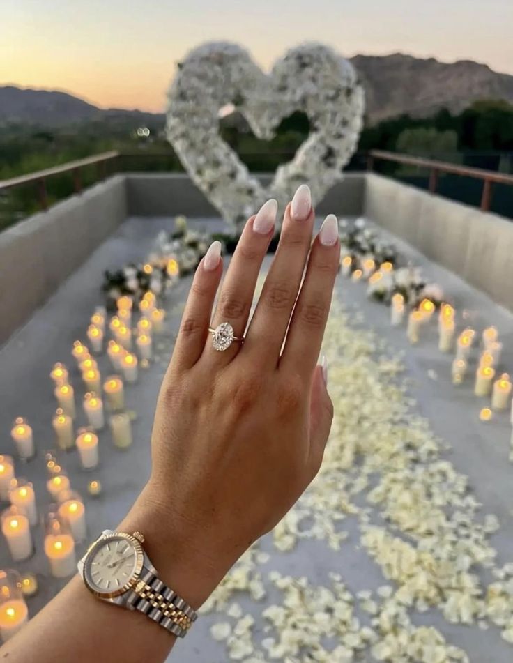 Romantic proposal setting with candles, white rose petals, and heart-shaped floral arch. Hand shows engagement ring and watch.
