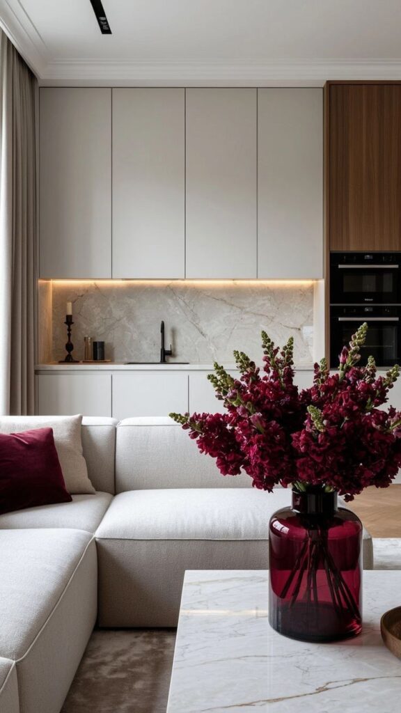 Modern living room with beige sofa, red flowers in a vase, marble coffee table, and sleek kitchen backdrop with under-cabinet lighting.