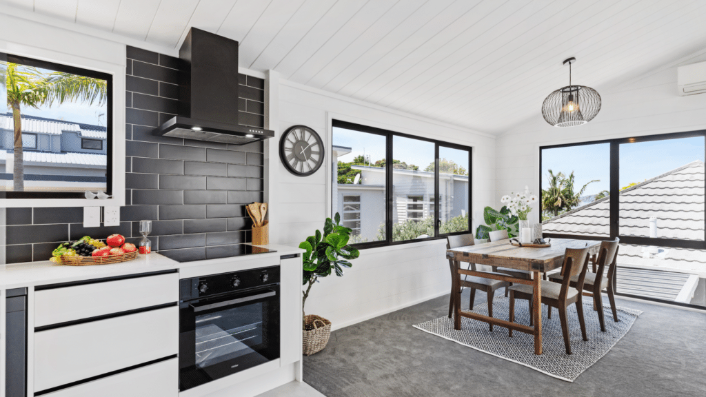 65fa72e549c7bbc502f2275c_Untitled design 9 Modern kitchen and dining area with black tiles, wooden table, and large windows offering views of trees and rooftops. | Sky Rye Design Modern kitchen and dining area with black tiles, wooden table, and large windows offering views of trees and rooftops.