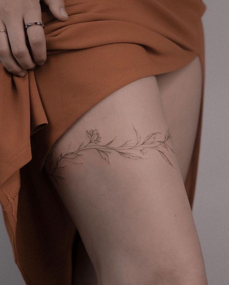_ 7 Delicate floral tattoo on a woman's thigh, partially covered by an orange dress, showcasing intricate leaves and buds. | Sky Rye Design Delicate floral tattoo on a woman's thigh, partially covered by an orange dress, showcasing intricate leaves and buds.