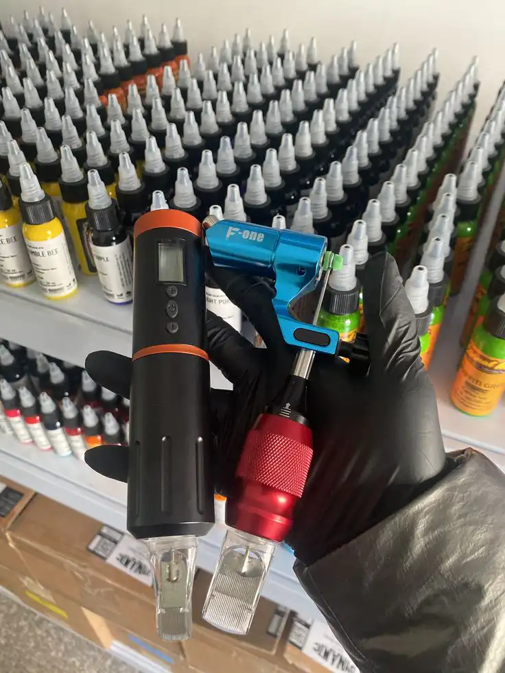Hand holding tattoo machine parts with shelves of ink bottles in the background, showcasing tattoo supplies in a studio setting.