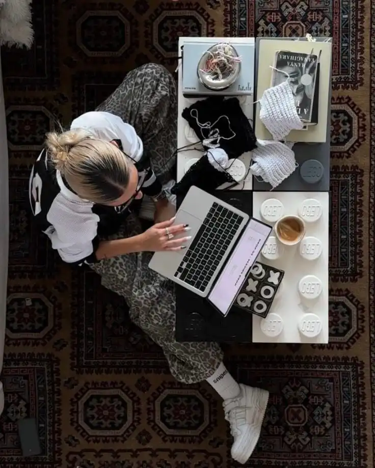 Person knitting at a laptop on a patterned rug with coffee, snacks, and books on the table beside. Cozy and creative workspace ambiance.