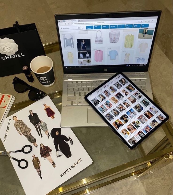 Fashion workspace with a laptop, tablet, Chanel coffee cup, and Saint Laurent cutouts on a gold table, showcasing fashion editing tools.