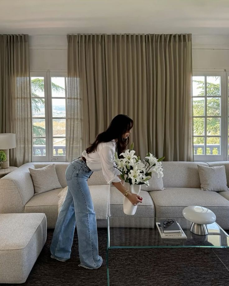 _ 78 Woman arranging lilies in a stylish living room with large windows, beige curtains, and modern furniture. Cozy and elegant home decor. | Sky Rye Design Woman arranging lilies in a stylish living room with large windows, beige curtains, and modern furniture. Cozy and elegant home decor.