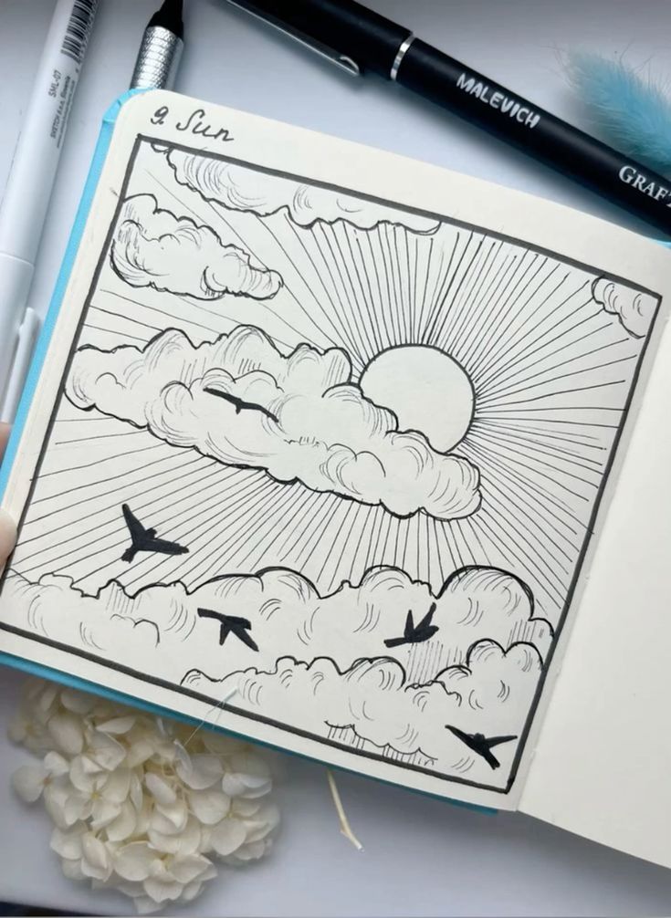 Sketch of sun, clouds, and birds in a notebook, with pens and flowers nearby. Hand-drawn illustration of a serene sky scene.
