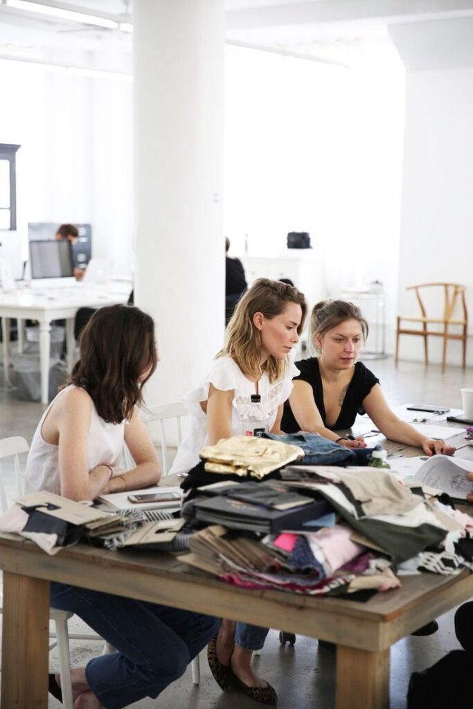 Women collaborating on fashion project in design studio, surrounded by fabric samples and creative materials.