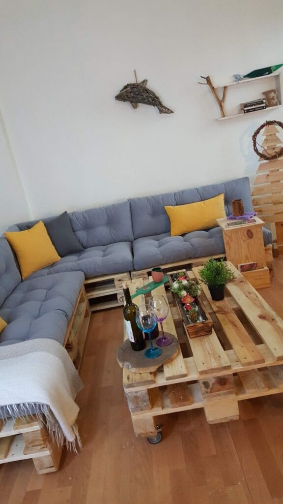 _ 8 Cozy living room with a blue pallet sofa, yellow cushions, wooden pallet table, wine, and decor on wooden floor. Rustic and inviting space. | Sky Rye Design Cozy living room with a blue pallet sofa, yellow cushions, wooden pallet table, wine, and decor on wooden floor. Rustic and inviting space.