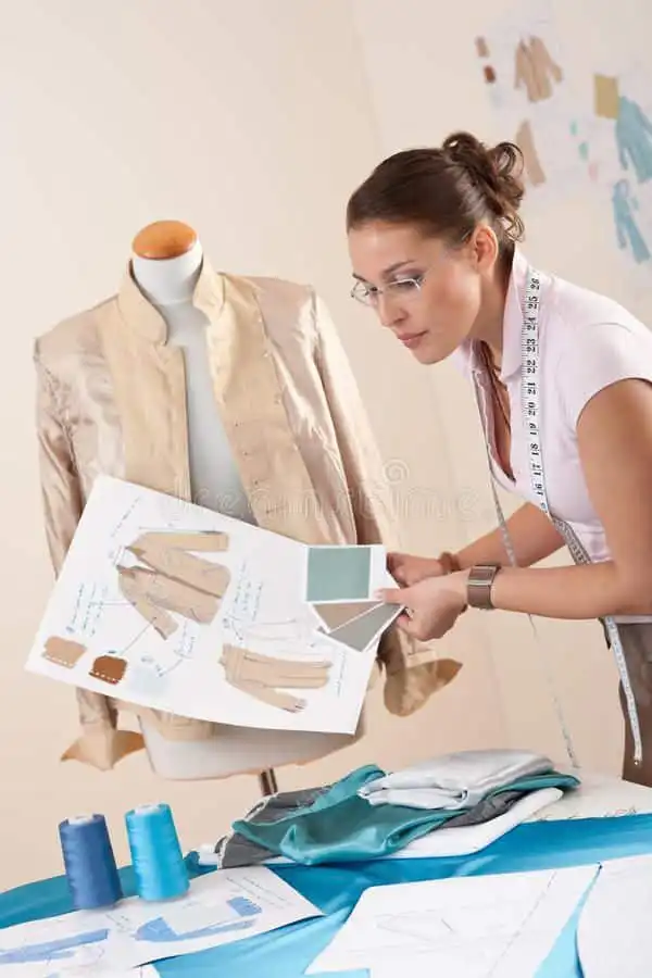 _ 8 Fashion designer reviewing jacket sketch with fabric swatches in studio. Mannequin in background with completed garment. | Sky Rye Design Fashion designer reviewing jacket sketch with fabric swatches in studio. Mannequin in background with completed garment.