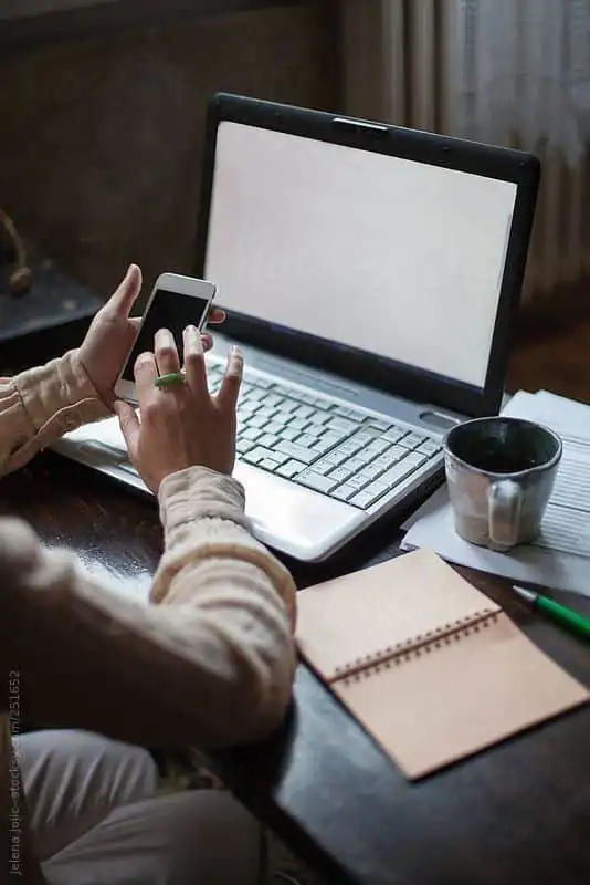 Person multitasks with phone and laptop at a desk, featuring a notebook and coffee mug, in a cozy home office setting.