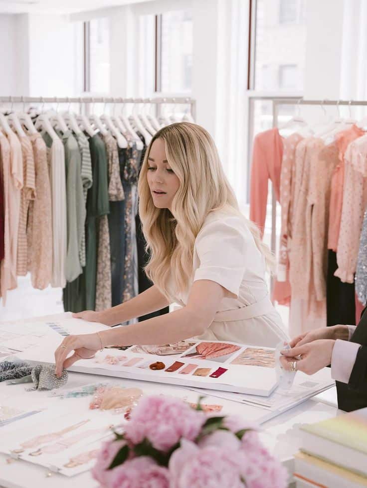 Fashion designer reviewing fabric swatches in a studio filled with colorful clothing racks and pink flowers in the foreground.