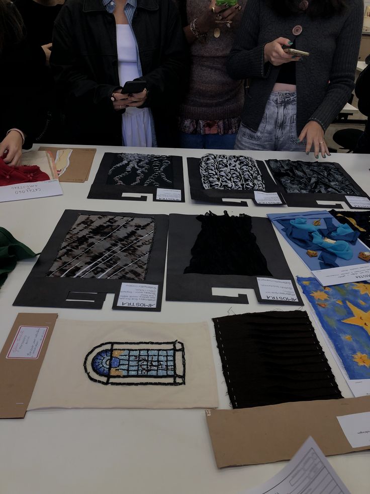 Textiles and fashion designs displayed on a table with people observing and taking photos.