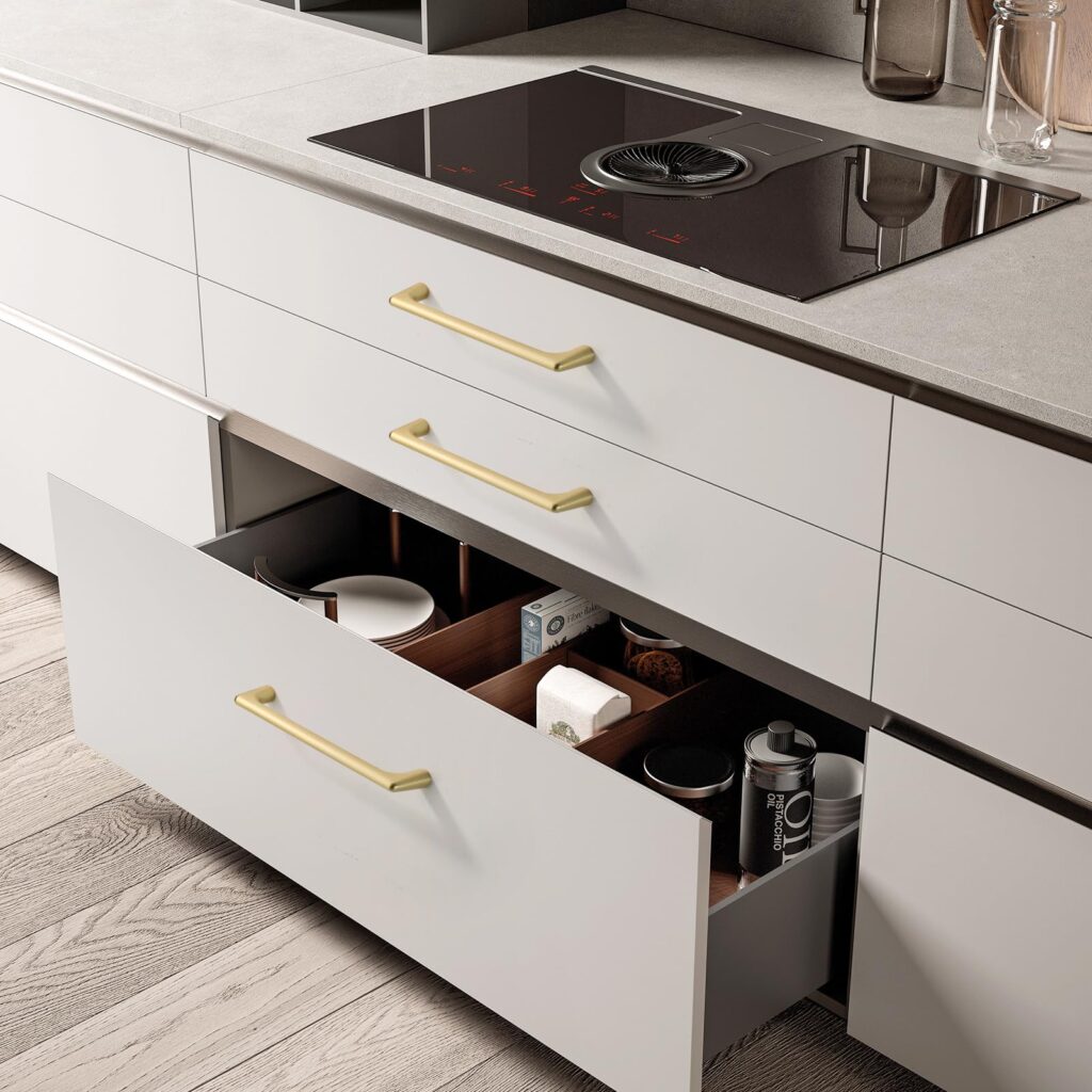 Modern kitchen with open drawers, gold handles, and induction cooktop, displaying organized cookware and spices on wood floor.
