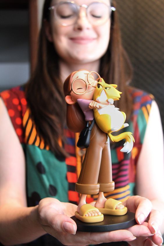Woman holding a cute cartoon figure, smiling with eyes closed. The character wears glasses and hugs a cat. Colorful background.