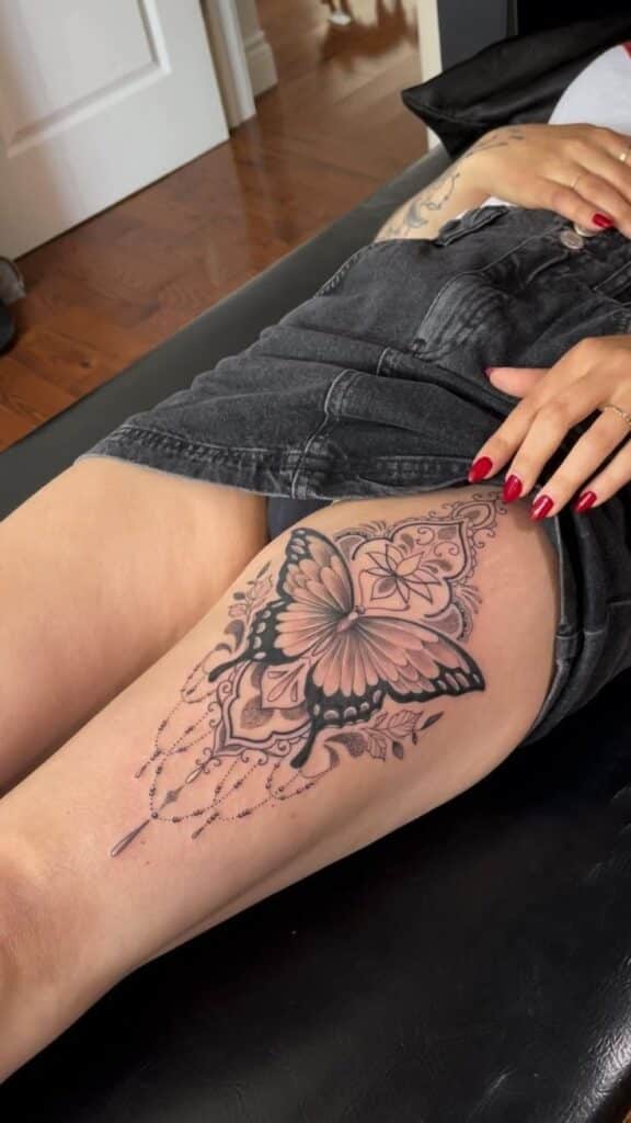 _ 9 Intricate butterfly thigh tattoo with floral and mandala elements on a person wearing black denim shorts, resting on a black surface. | Sky Rye Design Intricate butterfly thigh tattoo with floral and mandala elements on a person wearing black denim shorts, resting on a black surface.