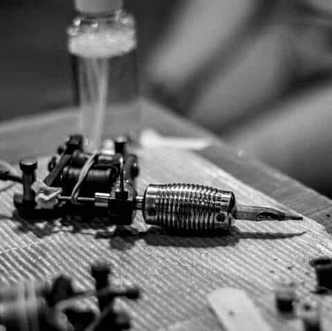 _ 9 Black and white image of a tattoo machine resting on a textured surface, with a bottle blurred in the background, capturing a tattoo studio vibe. | Sky Rye Design Black and white image of a tattoo machine resting on a textured surface, with a bottle blurred in the background, capturing a tattoo studio vibe.