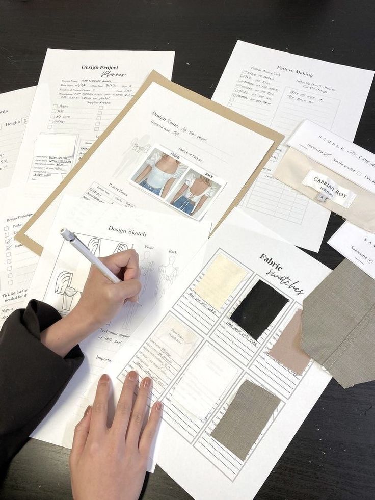 _ 9 A person sketching fashion designs surrounded by project plans, fabric swatches, and pattern-making sheets on a dark table. | Sky Rye Design A person sketching fashion designs surrounded by project plans, fabric swatches, and pattern-making sheets on a dark table.