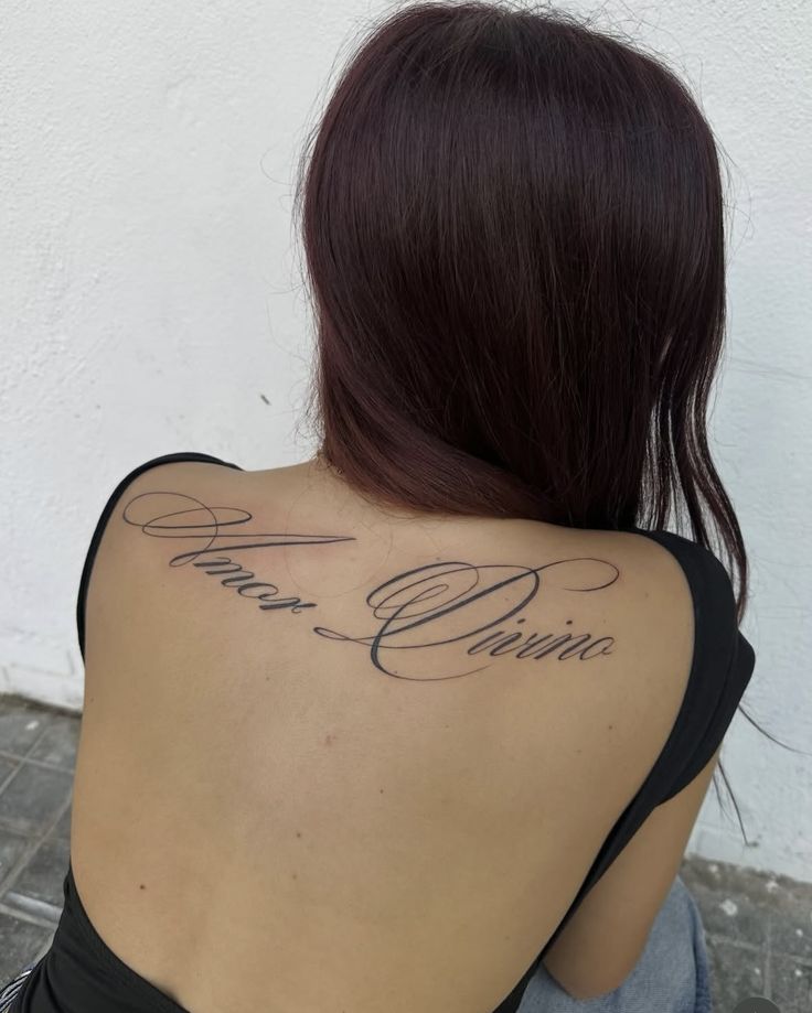 Screenshot Back tattoo reading Amor Divino in elegant script on a woman with long dark hair, wearing a black top, against a white background. | Sky Rye Design Back tattoo reading Amor Divino in elegant script on a woman with long dark hair, wearing a black top, against a white background.