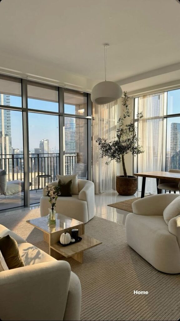 913a4168-281d-4d9f-861b-a91ea4221d20 Modern living room with large windows, cozy beige sofas, a marble table, and a potted plant. City skyline view, bright natural light. | Sky Rye Design Modern living room with large windows, cozy beige sofas, a marble table, and a potted plant. City skyline view, bright natural light.