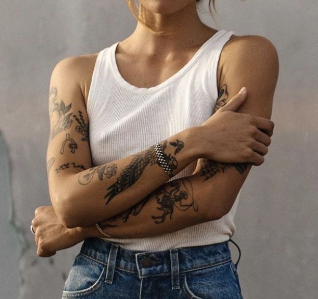 Person in a white tank top with tattooed arms crossed, wearing blue jeans and a silver bracelet.