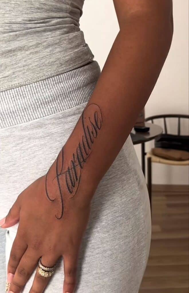 Close-up of a forearm tattoo reading Paradise in elegant script, on a person wearing gray clothing.