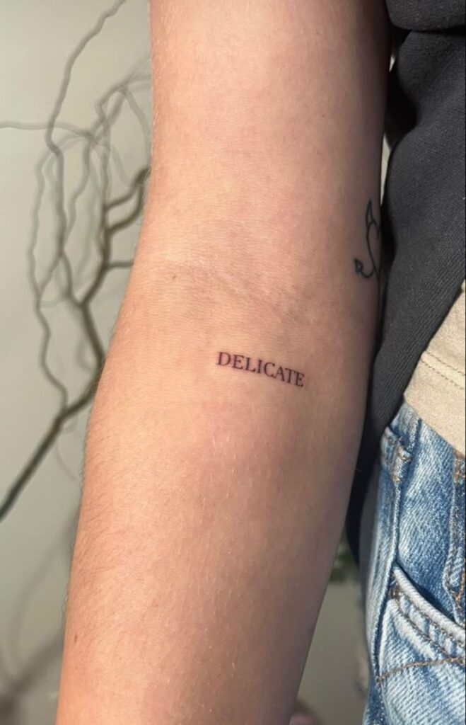 9c70b207-c206-45e5-bf73-d5784d0dbde2 Arm tattoo displaying the word Delicate in fine, black ink. Simple and minimal design on inner forearm against a soft background. | Sky Rye Design Arm tattoo displaying the word Delicate in fine, black ink. Simple and minimal design on inner forearm against a soft background. Best Fonts for Tattoos